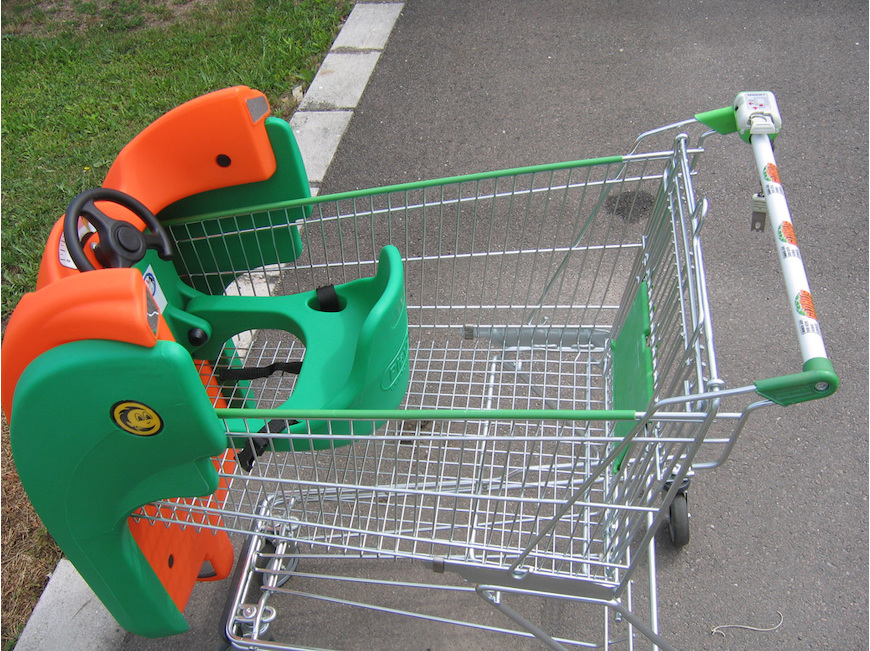 Na chodníku vedle travnaté plochy stojí nákupní vozík s připevněnou zelenou a oranžovou plastovou dětskou sedačkou, která je navržena tak, aby vypadala jako autíčko s volantem.