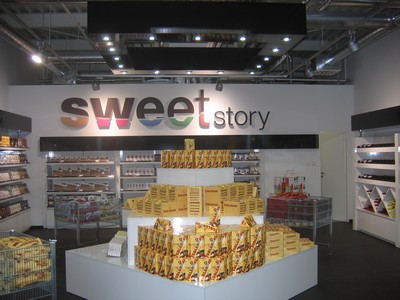 Cukrárna s názvem Sweet Story s jasným osvětlením, regály plné sladkostí a centrální vitrína zaplněná výrobky v žlutých obalech.